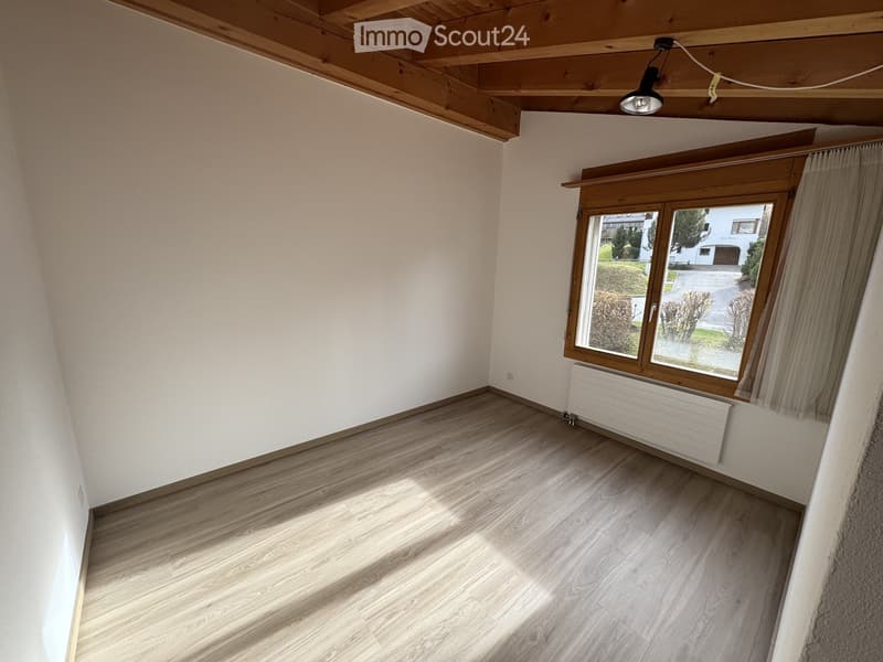 Empty room with wooden flooring, white walls, and a large window with wooden frame and curtains. Exposed wooden ceiling beams and a single ceiling light fixture are visible.