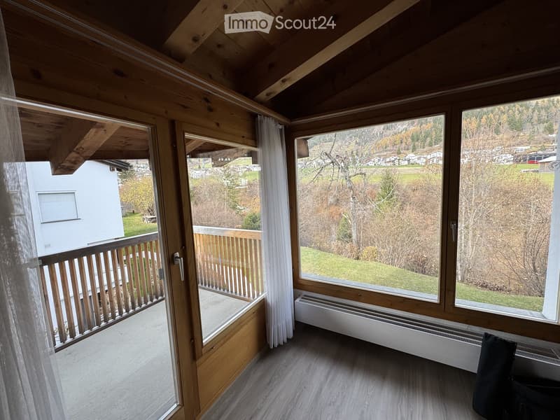 Bright room with wooden beams, large windows, and a view of the surrounding landscape. Features a balcony with wooden railings and a radiator under the windows.