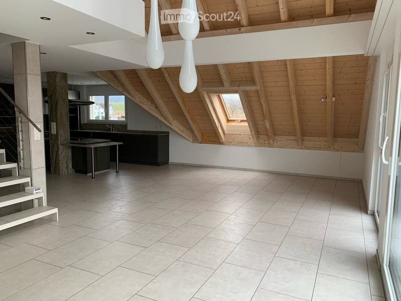 Open-plan space with a modern kitchen area featuring a central island, cabinets, and pendant lights, connected to a tiled living area with a staircase leading upwards.