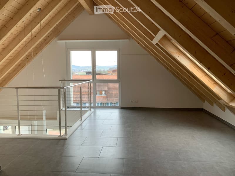 Empty attic space with wooden slanted ceilings, large windows, and a modern glass railing leading to a balcony.