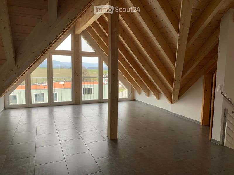 Bright, spacious attic room with wooden beams, large windows, and a balcony overlooking a green landscape.