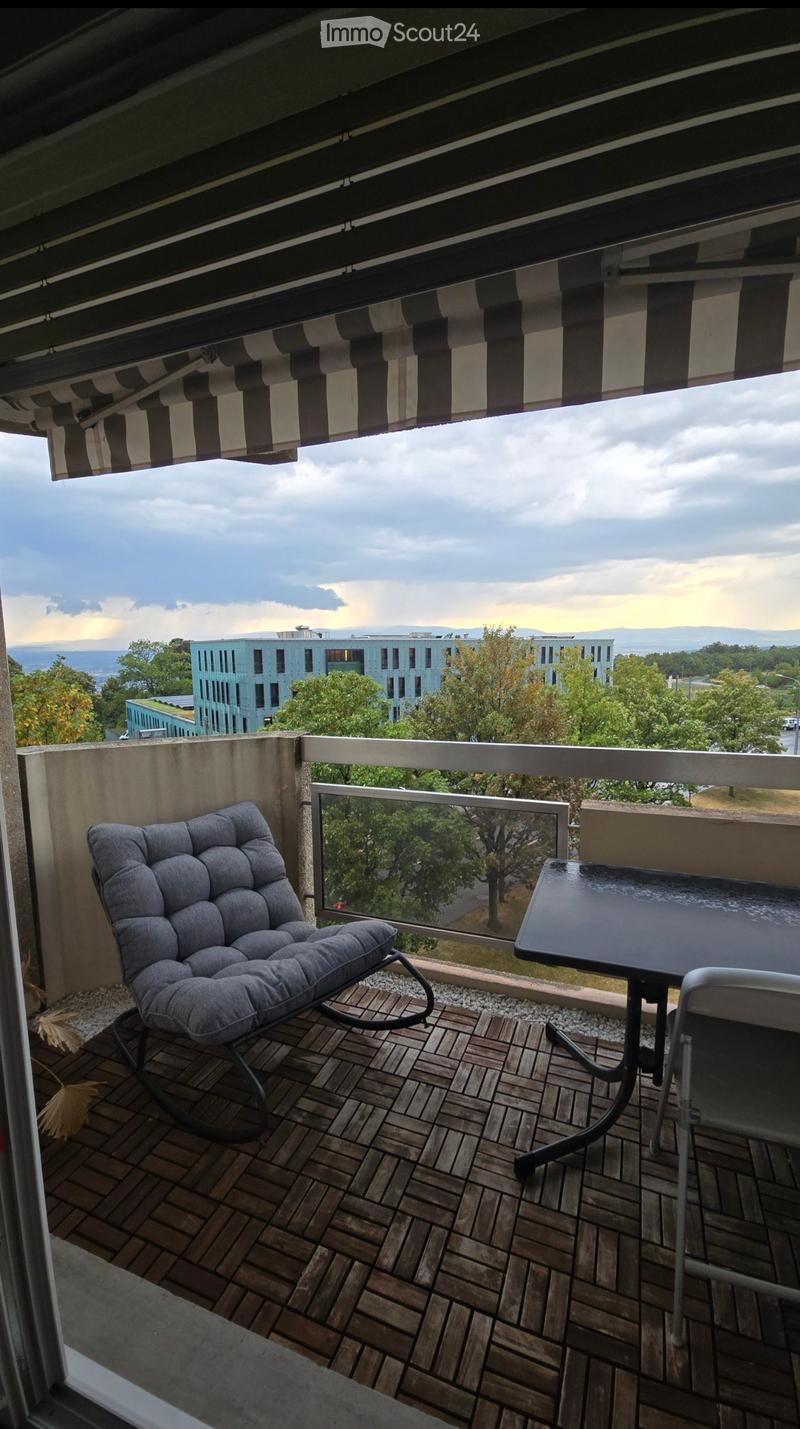Balcony with wooden decking, grey cushioned chair, black table, and a view of the city and mountains.