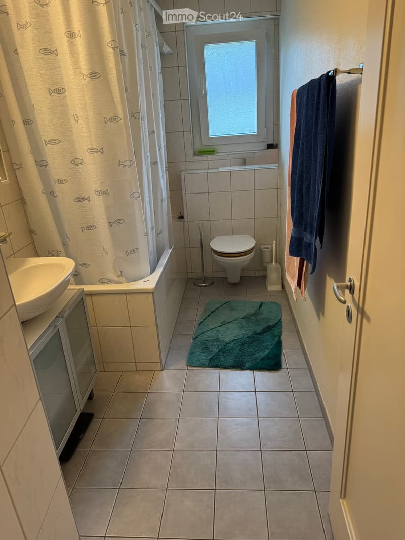 Bathroom with tiled walls and floor, bathtub with fish-patterned shower curtain, toilet, sink, mirror, and towels on a rack.