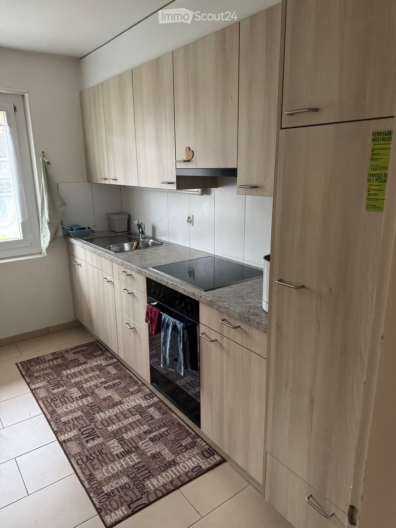 Modern kitchen with light wood cabinets, stainless steel sink, electric stove, and oven. Includes a decorative rug with coffee-themed text.