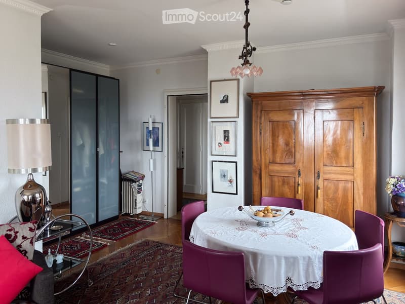 Elegant dining area with a white tablecloth-covered table, purple chairs, a wooden cabinet, and decorative lamps. The room features framed artwork on the walls and a cozy seating area with a sofa and decorative pillows.