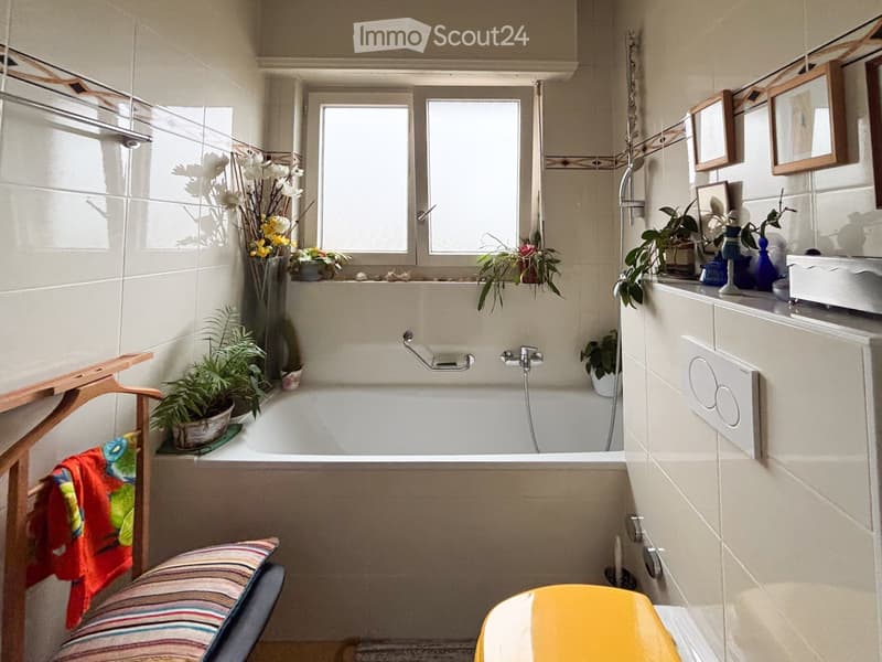 Bathroom with a bathtub, window, potted plants, framed pictures, a yellow chair, and a striped cushion on a chair.