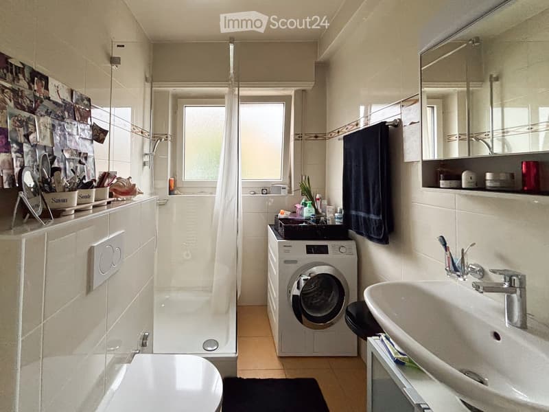 Modern bathroom with a toilet, sink, mirror, shower, and washing machine. Features a towel, toothbrush holder, and various toiletries on the sink counter.