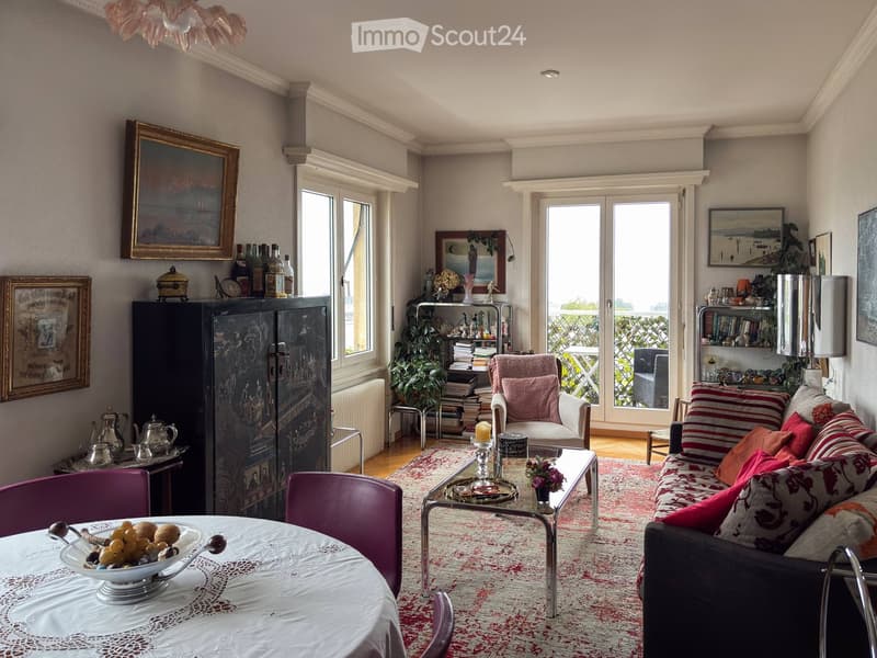 Elegant living room with a patterned rug, a sofa with colorful cushions, a glass coffee table, a dining table with purple chairs, a cabinet, and a bookshelf. Large windows and a balcony are visible.