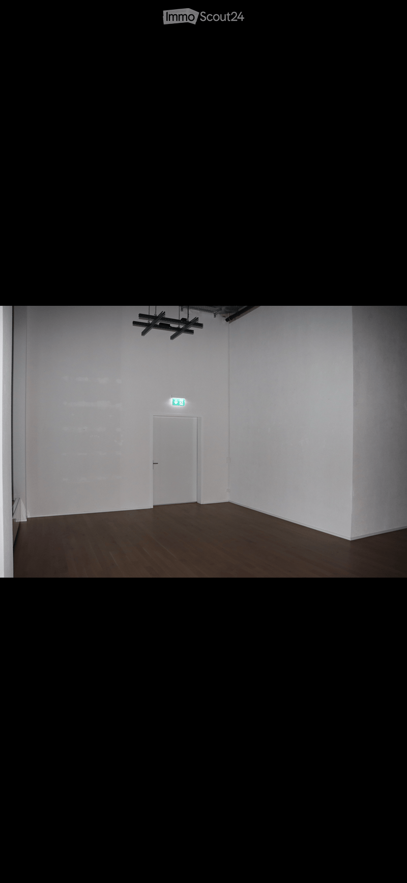Empty room with white walls, wooden floor, and a single white door with an emergency exit sign above it.