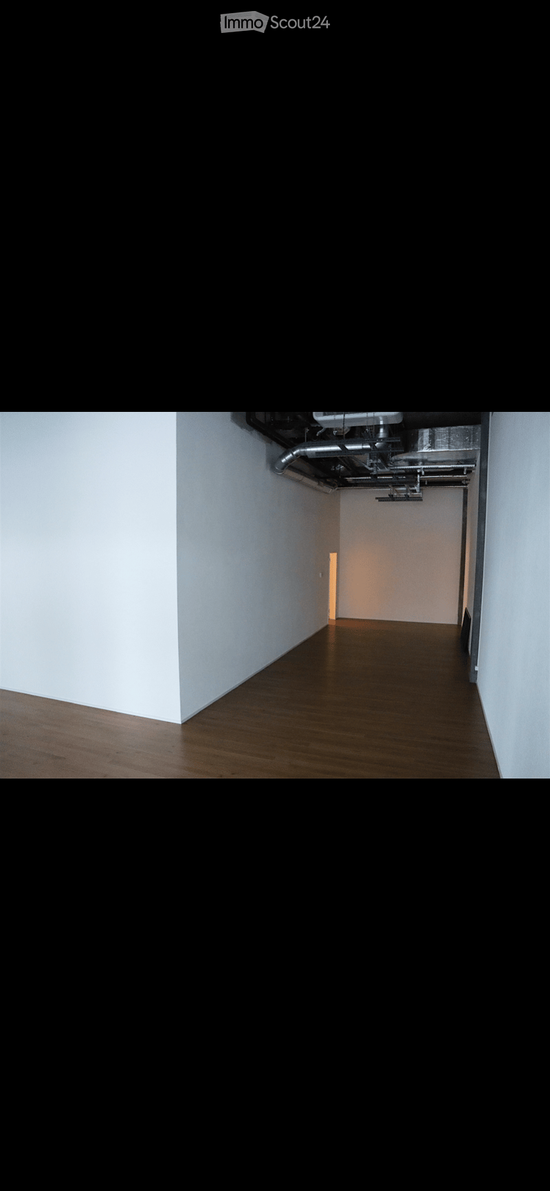 Empty hallway with wooden flooring and white walls, leading to a room with a visible door and a small light fixture.