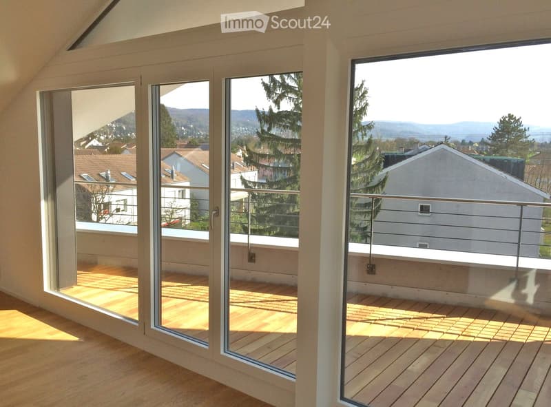 Large, open balcony with wooden decking and modern glass railings, offering scenic views of the surrounding area and distant hills.