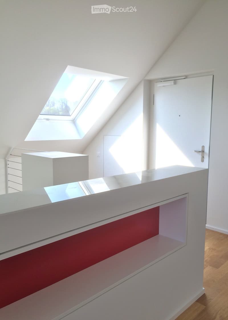 Bright, modern attic space with wooden flooring, white walls, and skylight windows allowing natural light. Features a white door and a sleek white counter with a red accent panel.