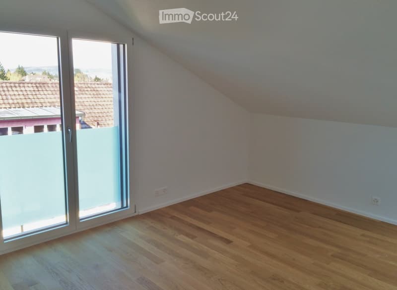 Empty attic room with wooden flooring, white walls, and large sliding glass doors leading to a balcony with a view of the surrounding area.