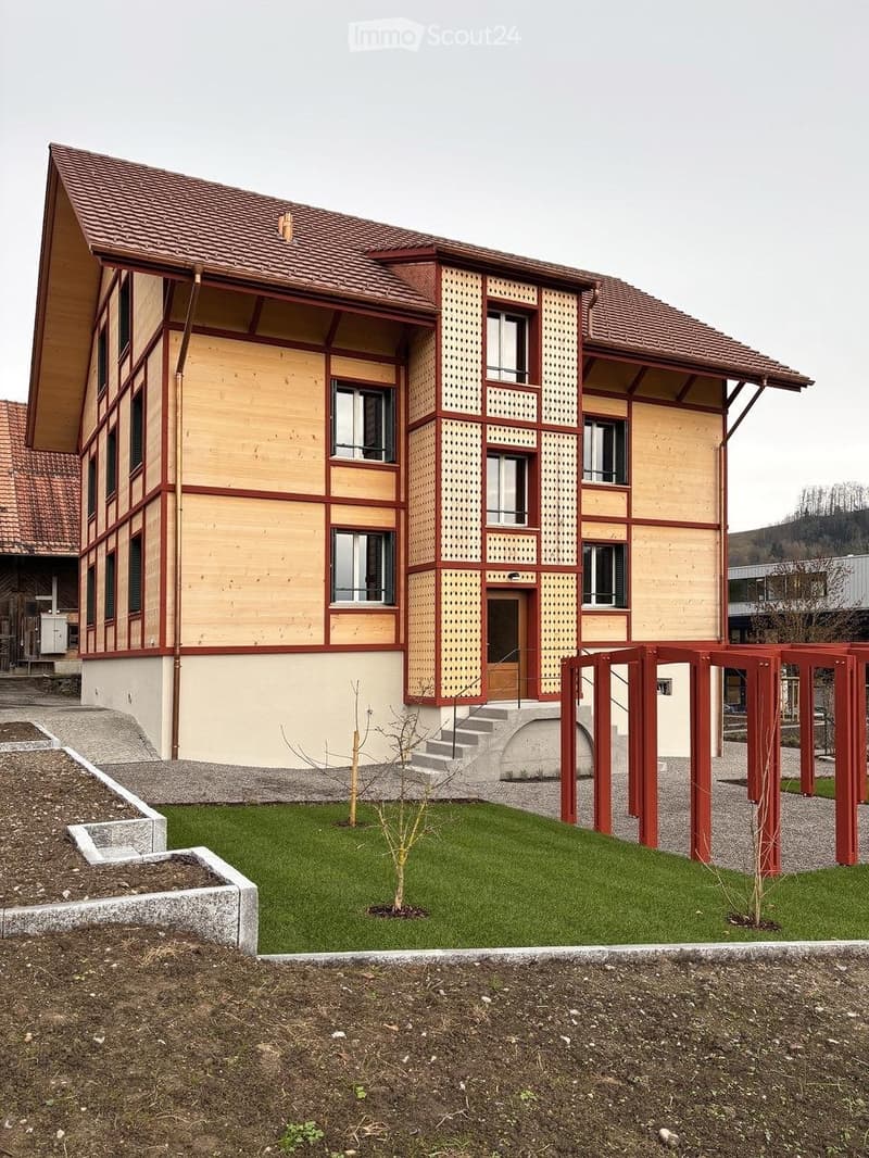 Two-story building with a red-tiled roof, wooden facade, and multiple windows. The entrance features a small staircase leading to a door, with a landscaped lawn and young trees in the foreground.