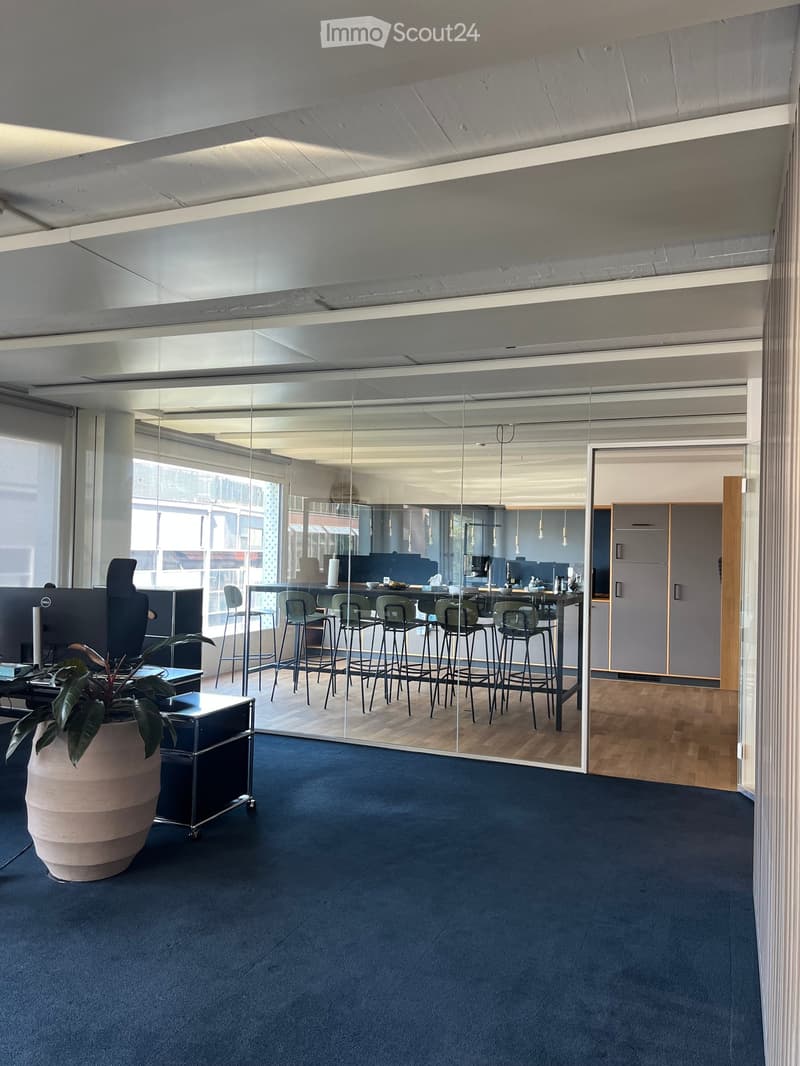 Modern office space with blue carpet, glass partition, stool chairs around a table, a large screen, and a potted plant.
