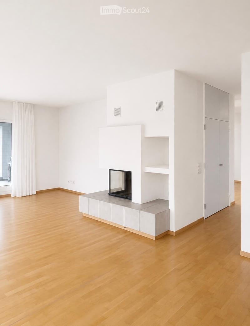 Empty room with light wooden flooring, white walls, and a built-in fireplace with a mirror above it. There are built-in shelves and cabinets, and a large window with white curtains on the left side.