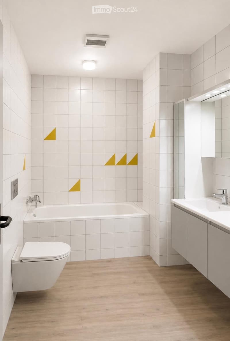 Modern bathroom with white tiled walls, wooden floor, bathtub, toilet, sink, and mirror. Features geometric yellow wall decals and sleek fixtures.