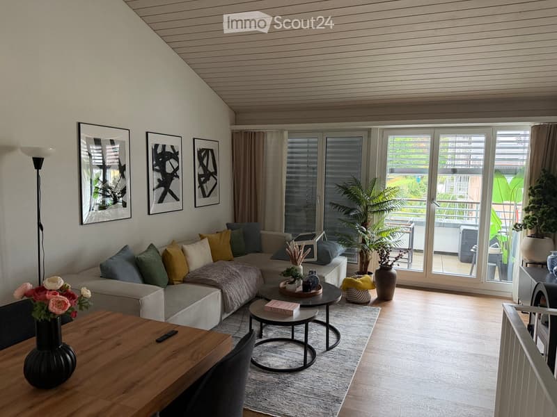 Modern living room with a grey sectional sofa, colorful cushions, a wooden coffee table, side tables, vases, and a floor lamp. Large windows and sliding doors lead to a balcony with outdoor furniture.
