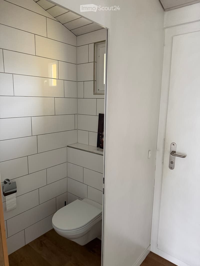 Modern, compact bathroom with white tiled walls, a toilet, a toilet paper holder, and a small mirror above a sink.