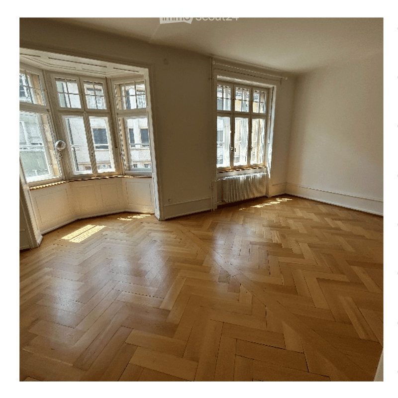Empty room with wooden herringbone flooring, large windows, and radiators beneath them.