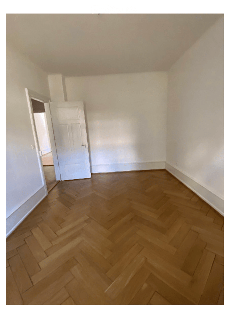 Empty room with wooden herringbone flooring, white walls, and a white door leading to another space.