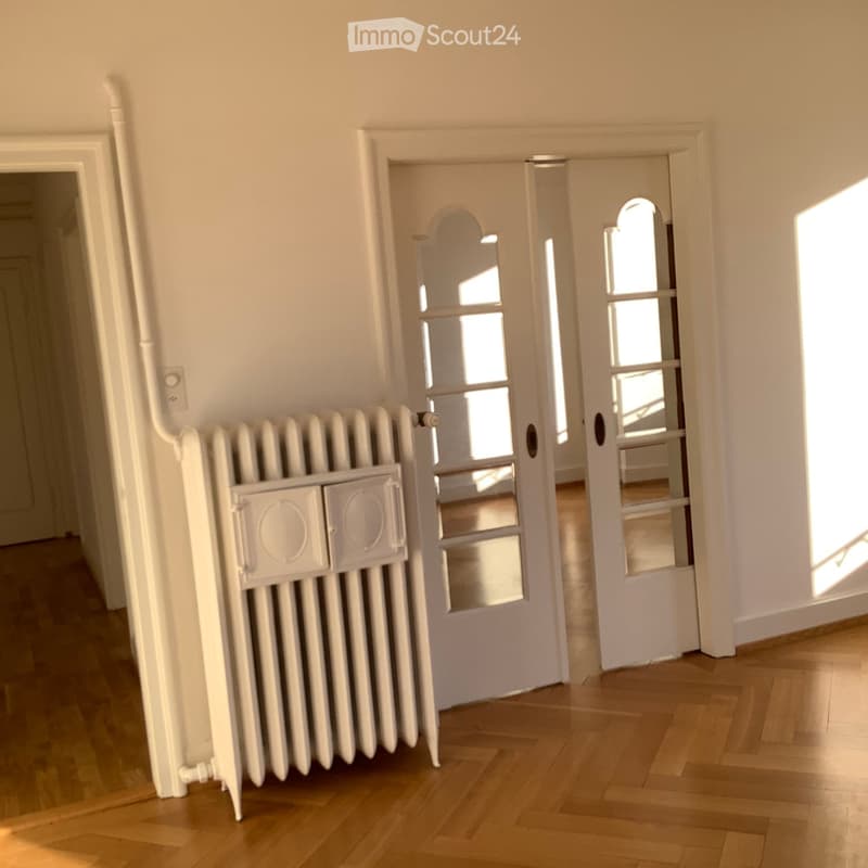 Empty hallway with wooden flooring, white walls, radiator, and two open doors leading to other rooms.