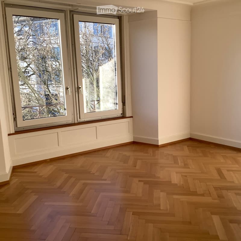 Empty room with wooden herringbone flooring, white walls, and large windows with white frames and a radiator cover below them.