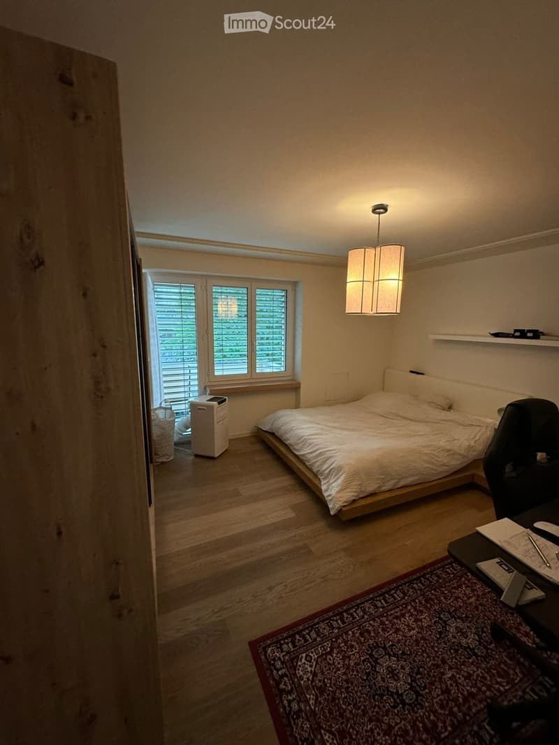 Cozy bedroom with wooden flooring, a bed with white bedding, a desk with a chair, a patterned rug, and a hanging lamp.