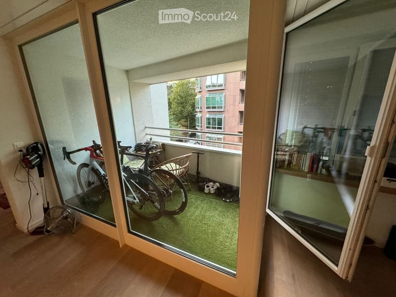 Balcony with green artificial turf, two bicycles, a small table, a chair, and a fan. The balcony has large glass doors leading to the interior.