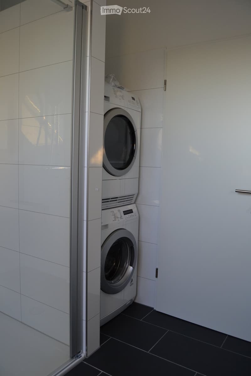 A compact laundry area with a stacked washing machine and dryer unit, white tiled walls, and dark tiled flooring.
