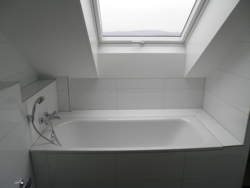 Modern bathroom with a white bathtub, chrome faucet, and handheld showerhead. The room features white tiled walls and a skylight window providing natural light.
