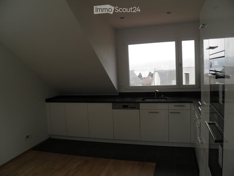 Modern kitchen with white cabinets, black countertops, and a stainless steel sink. The room features large windows with a view of rooftops and a slanted ceiling.