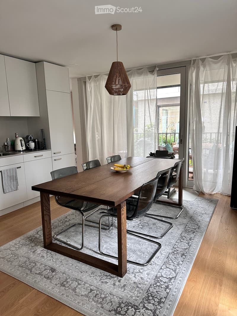 Modern dining area with a large wooden dining table surrounded by chairs, a kitchen area with white cabinets and appliances, and large windows with sheer curtains leading to a balcony.