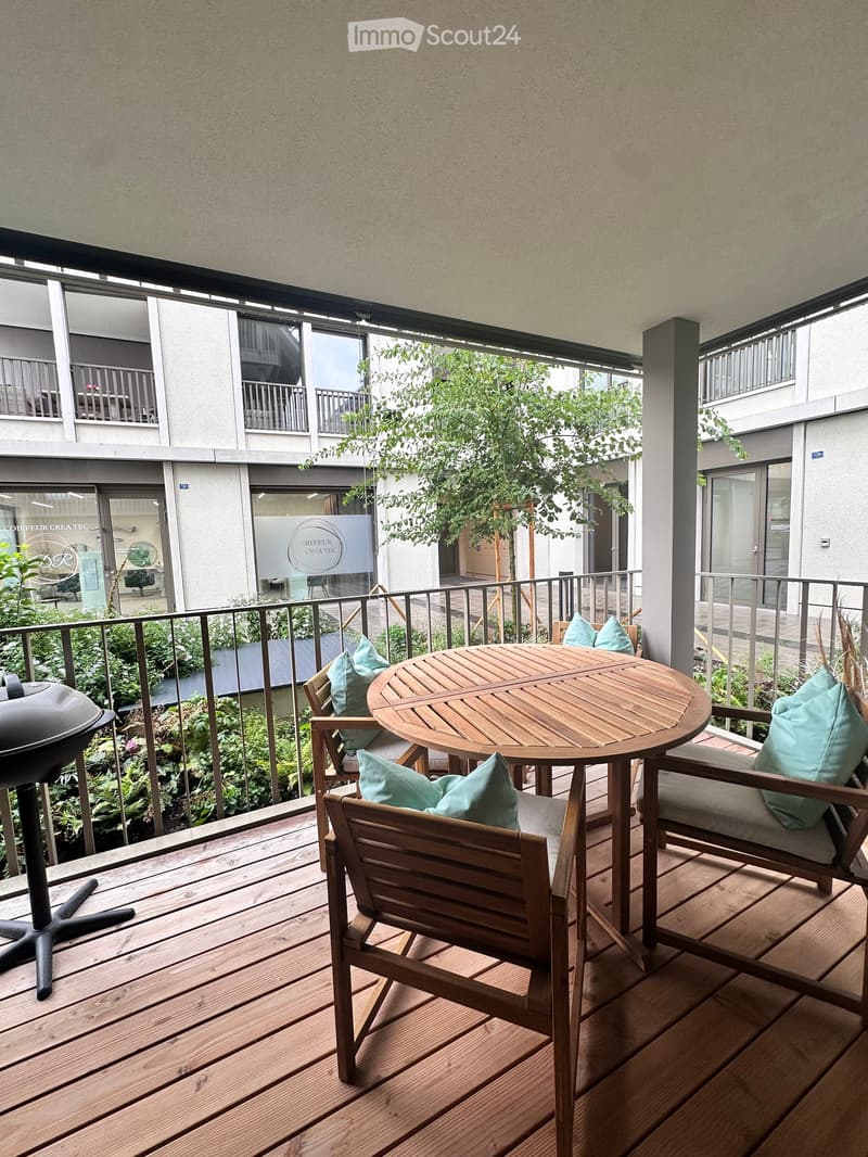 Wooden balcony with a round table, four chairs, and teal cushions, surrounded by greenery and a railing.