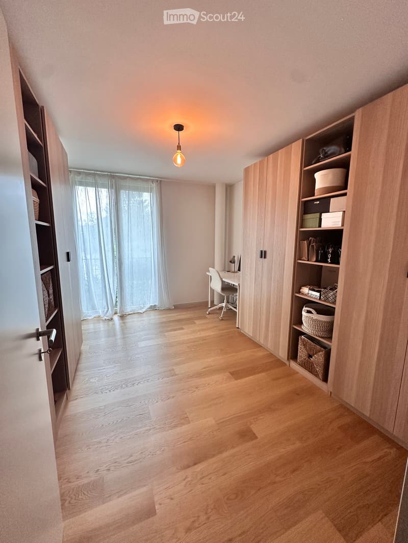 A modern, well-lit room with wooden flooring, a desk with a chair, built-in cabinets, and a large window with sheer curtains.