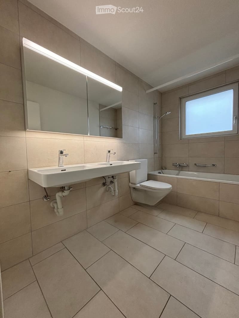 Modern bathroom with beige tiles, a double sink, toilet, bathtub, and large mirror with built-in lighting.
