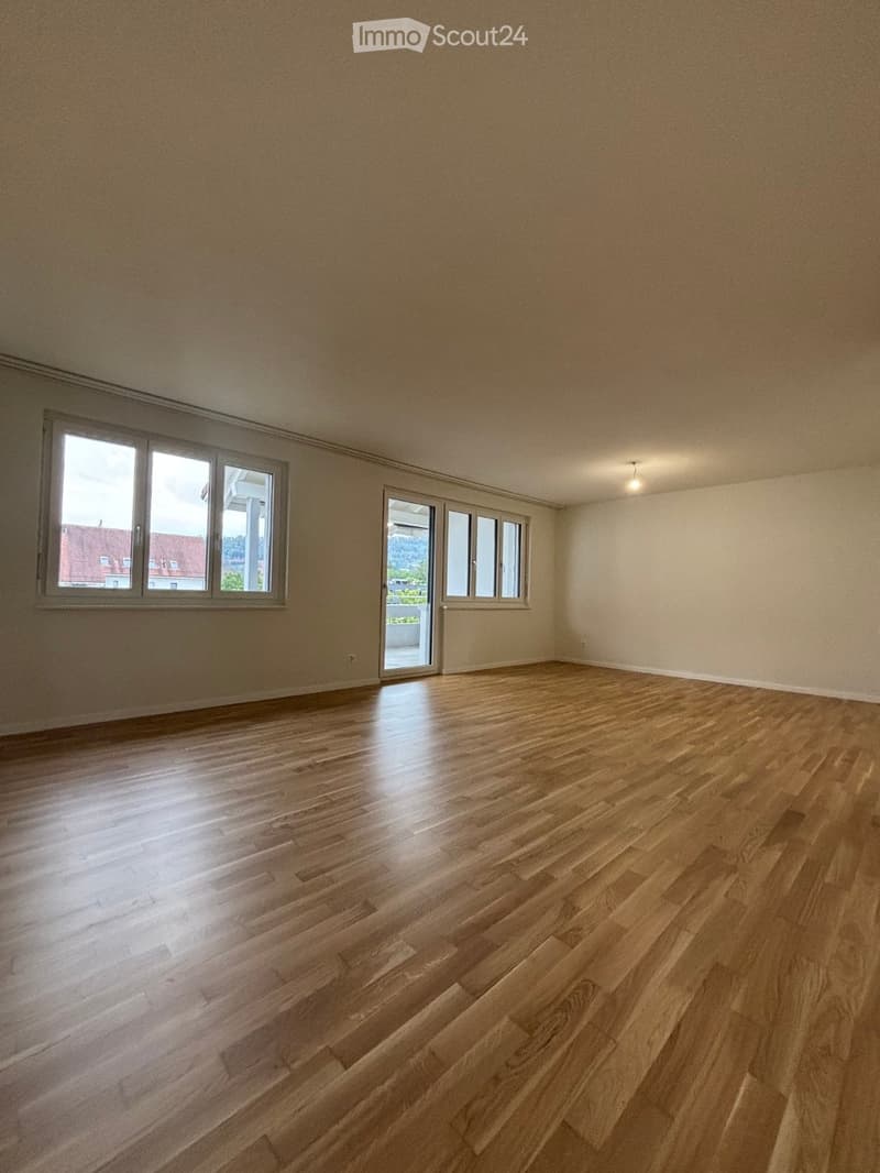 Empty room with wooden flooring, large windows, and a sliding glass door leading to a balcony.