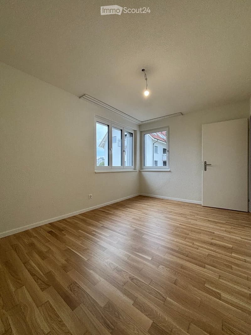 Empty room with wooden flooring, white walls, and two windows offering a view of neighboring buildings.