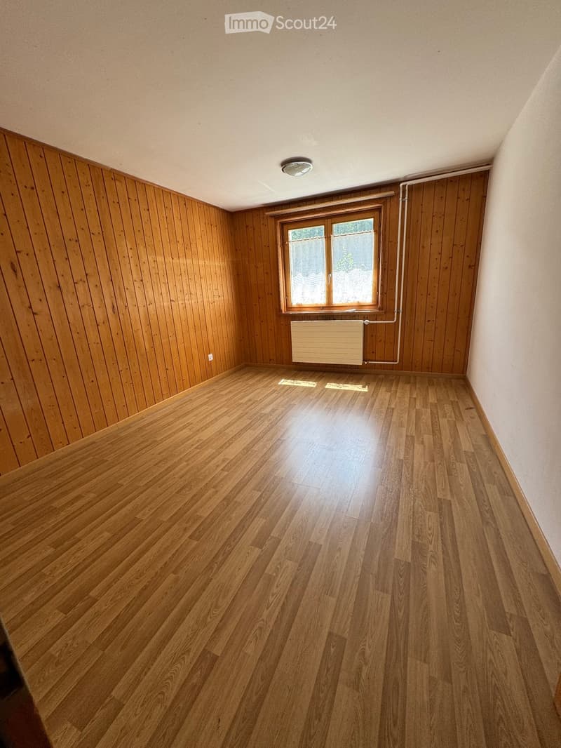 Empty room with wooden walls and floor, a window, radiator, and ceiling light fixture.