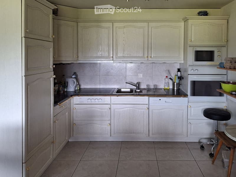A modern kitchen with light-colored cabinets, a stainless steel sink, a microwave, an oven, and a small table with a stool.