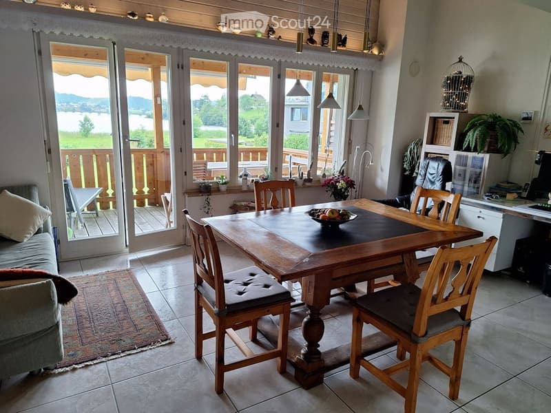 Bright living and dining area with large windows and a view of the lake. Features a sofa, dining table with chairs, a birdcage, and a cabinet with a laptop.
