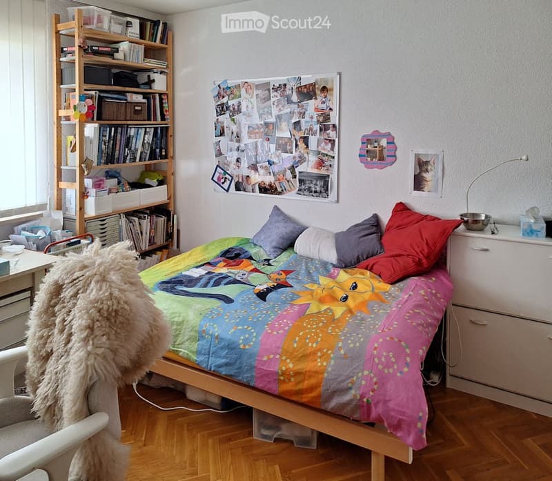 Cozy bedroom with a colorful bedspread, wooden floor, bookshelf, and personal decorations including photos and artwork.