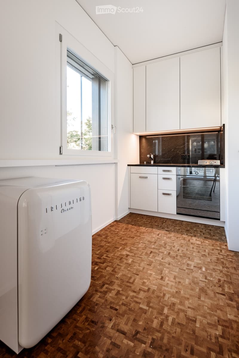 Modern kitchen with white cabinets, wooden herringbone flooring, stainless steel appliances, and a large window.