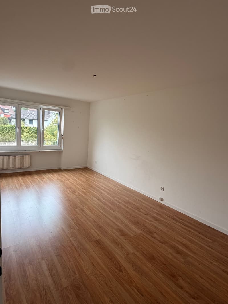 Empty room with light wooden flooring, white walls, and large windows with white frames. A radiator is visible under the windows on the left side.