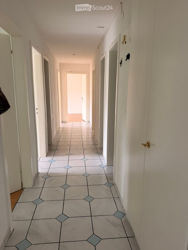 Empty hallway with white tiled floor and light-colored walls, featuring multiple doors and a lamp on the left side.