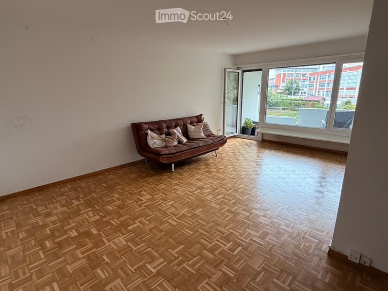 Living room with wooden herringbone flooring, a brown leather sofa with colorful pillows, and large windows opening to a balcony.