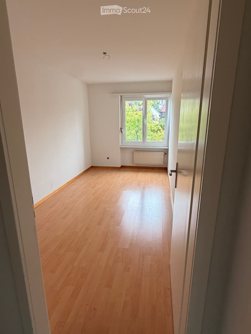 Empty room with wooden flooring, white walls, and a large window with a view of greenery.