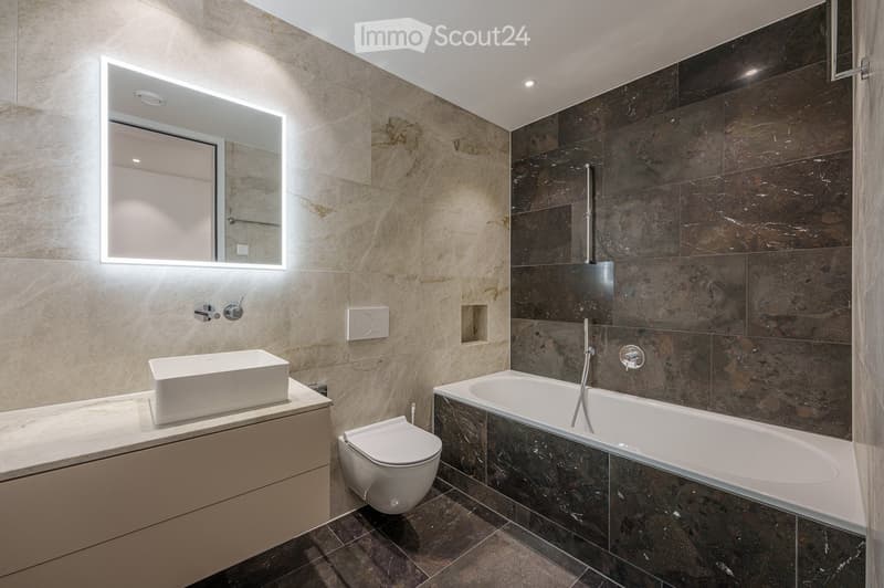 Modern bathroom with marble tiles, a white sink, bathtub, toilet, and illuminated mirror.