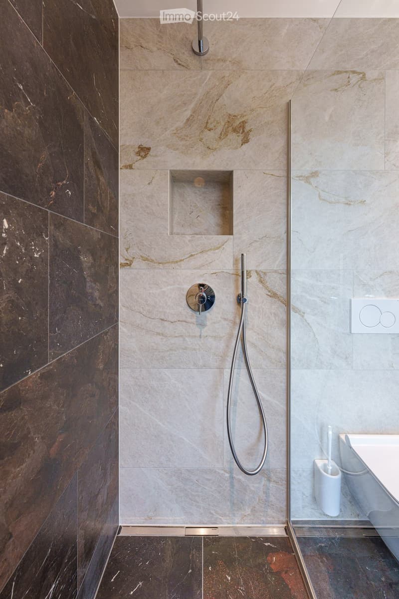 Modern bathroom with marble tiles, a glass-enclosed shower area, a sleek showerhead, and a modern faucet. A toothbrush holder is visible near the toilet.