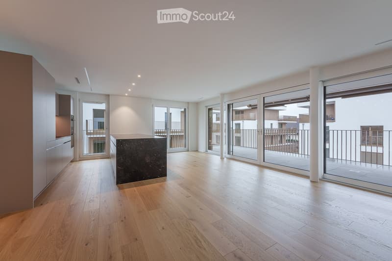 Bright, open-plan room with wooden flooring, large glass sliding doors leading to a balcony, and modern built-in kitchen units on one side.